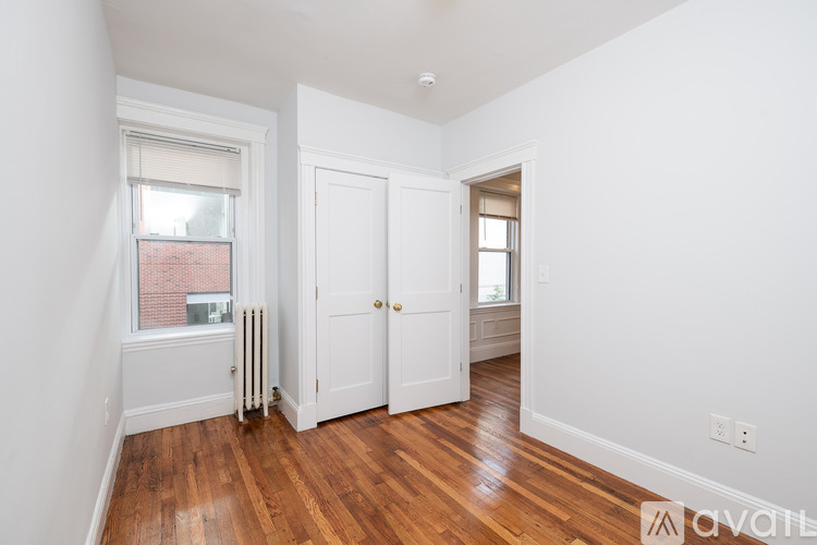A room with white walls and wooden floors, with a window showing a red building outside.