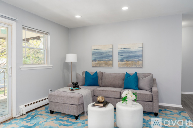 A living room with a grey couch, a small ottoman, a table with a vase of flowers, and two paintings on the wall.