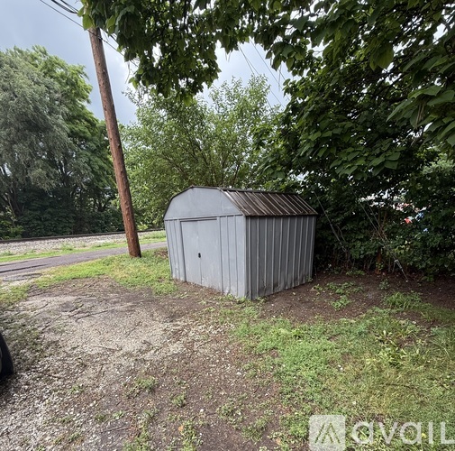 A grey metal shed sits in a grassy area with trees and a telephone pole nearby.