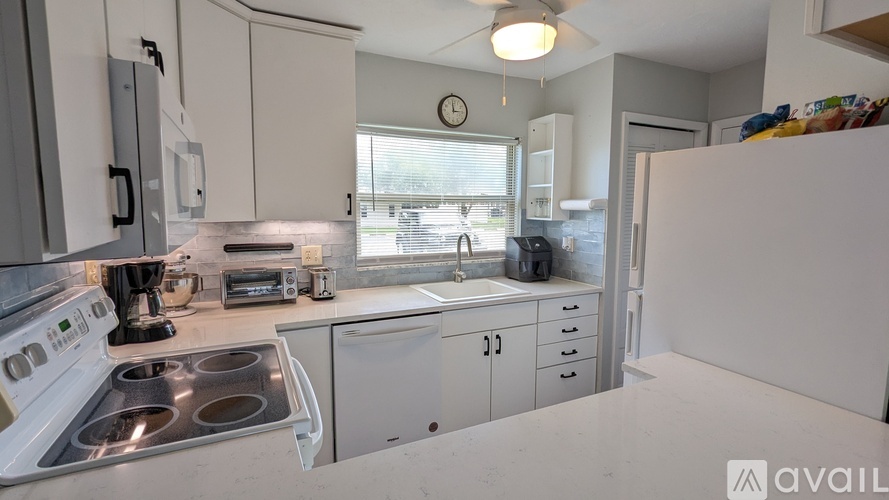 A kitchen with white cabinets and appliances, a clock on the wall, and a window with blinds.