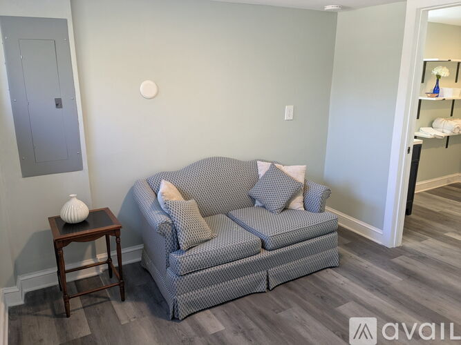 A grey couch with pillows sits in a room with a grey wall and a wooden table with a white vase on it.