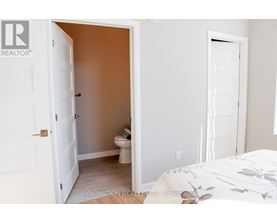 A white door is open to a small bathroom with a toilet and a bedroom with a white bed.