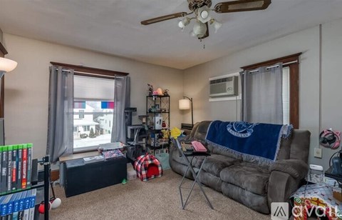 A living room with a grey couch, a fan, a window with curtains, and a bookshelf.