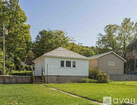 A small house with a white exterior and a brown roof is surrounded by a grassy area and trees.