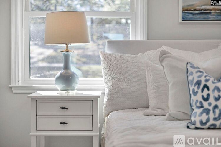 A bedroom with a white bed, a nightstand with a lamp, and a painting on the wall.