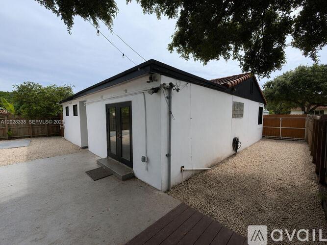 A small white building with a black roof and a black door is surrounded by a gravel driveway.