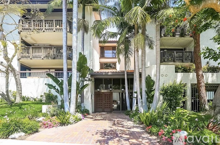 A white building with a balcony is surrounded by green plants and trees.