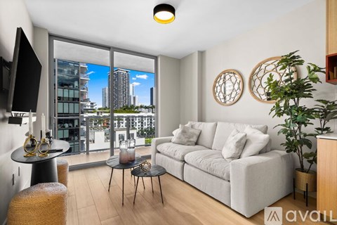 A living room with a couch, a table, a chair, a plant, and a window overlooking a cityscape.