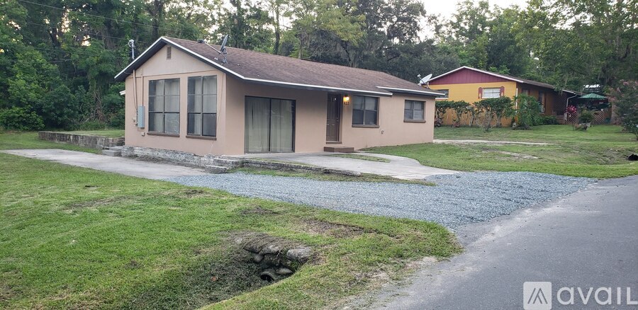 A house with a brown roof and a beige exterior is surrounded by a gravel driveway and a grassy lawn.
