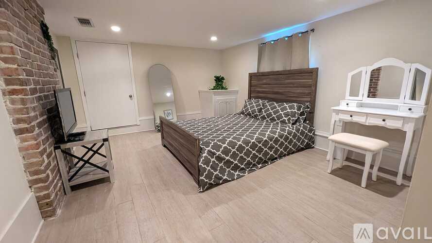 A bedroom with a brick wall, a bed with a black and white comforter, a white chair, and a small table with a plant on it.