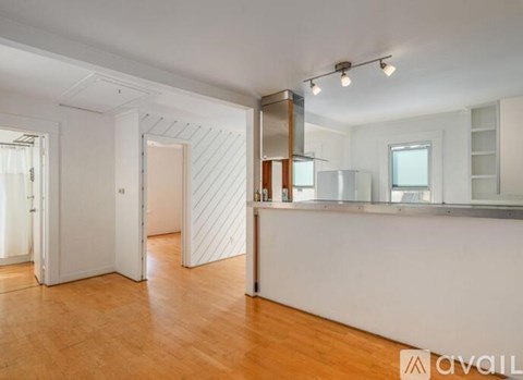 A spacious room with wooden floors and white walls, featuring a bar area with a counter and a refrigerator.