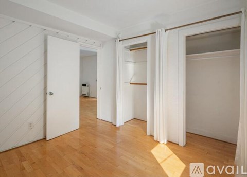 A room with wooden floors and white walls, with a door on the left and a window with curtains on the right.