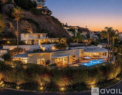 A large house with a pool is surrounded by a hedge and lit up at dusk.