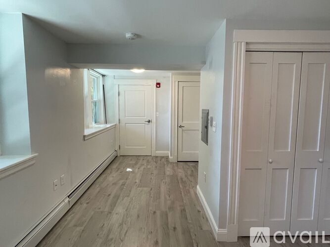 A room with a wooden floor and white walls, with a door on the right and a window on the left.