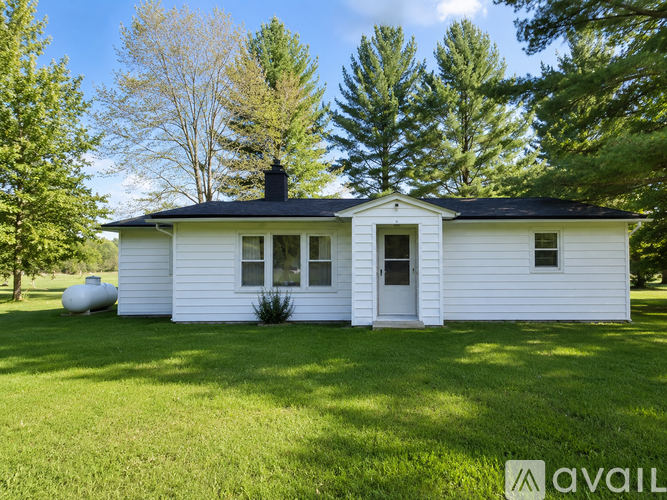 A white house with a black roof and a chimney is surrounded by green grass and trees.