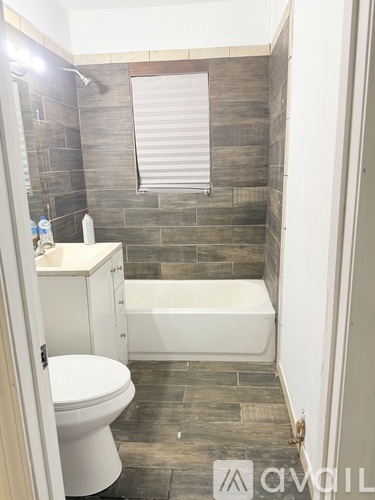 A white toilet sits in a bathroom with a white bathtub and a window with blinds.