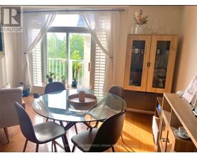 A glass dining table with four chairs is in the foreground of a well-lit room with a balcony outside.
