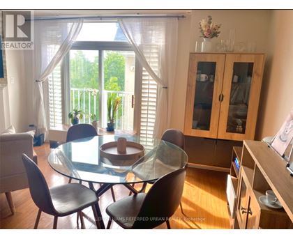 A glass dining table with four chairs is in the foreground of a well-lit room with a balcony outside.