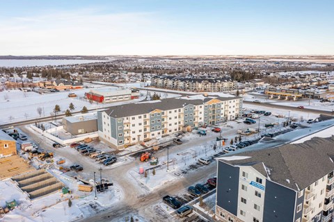 A view of a snowy parking lot with several cars and buildings in the background.