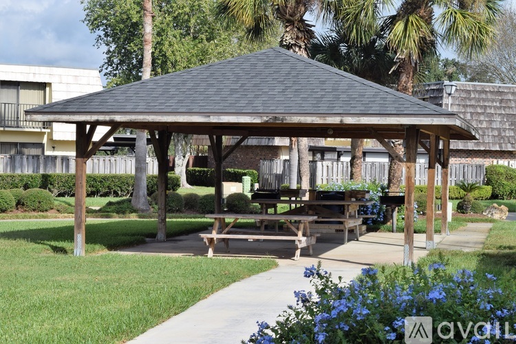 A pavilion with a wooden roof and a picnic table in front of a fence with a walkway and trees in the background.