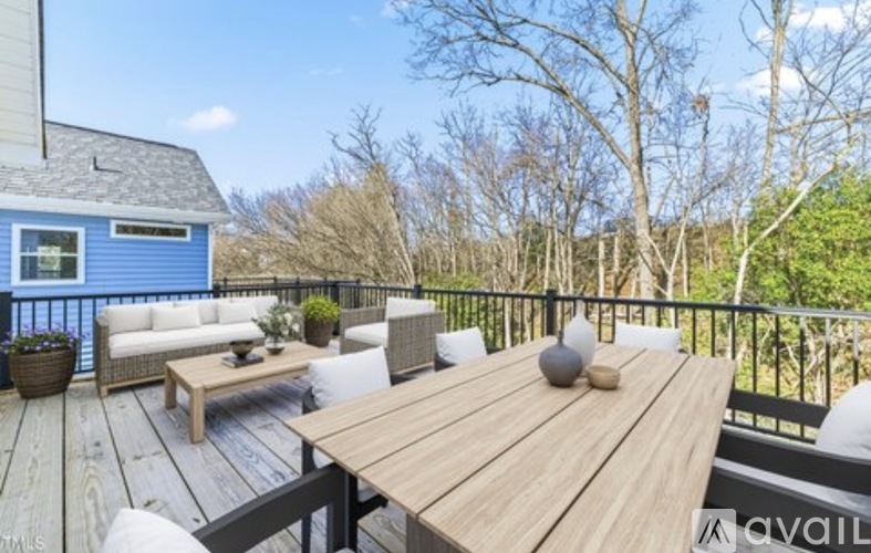 A wooden table with a vase on it sits on a deck with a blue house and trees in the background.