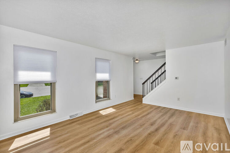 A spacious room with wooden floors and white walls, featuring two windows and a staircase.