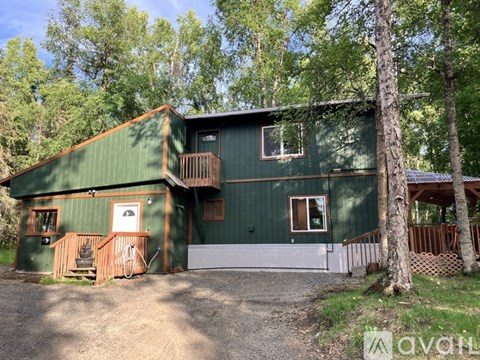 A green wooden cabin with a white door and windows is surrounded by trees.