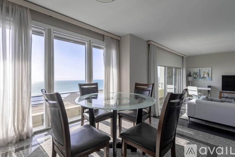 A modern dining room with a glass table and chairs, a sofa, and a view of the ocean through the windows.