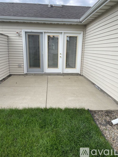 A patio with a door and windows is surrounded by a concrete floor and a grassy area.