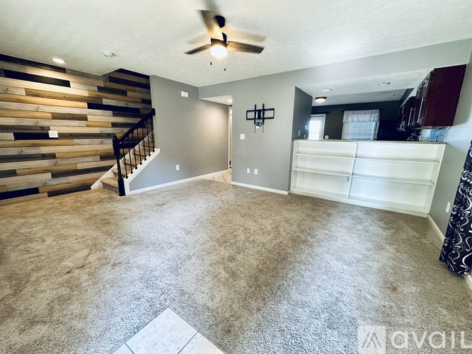A spacious room with a carpeted floor, a ceiling fan, and a wooden staircase.