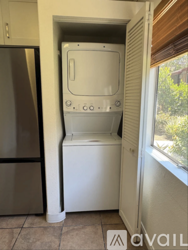 A small white washing machine is in a narrow space between a refrigerator and a window.