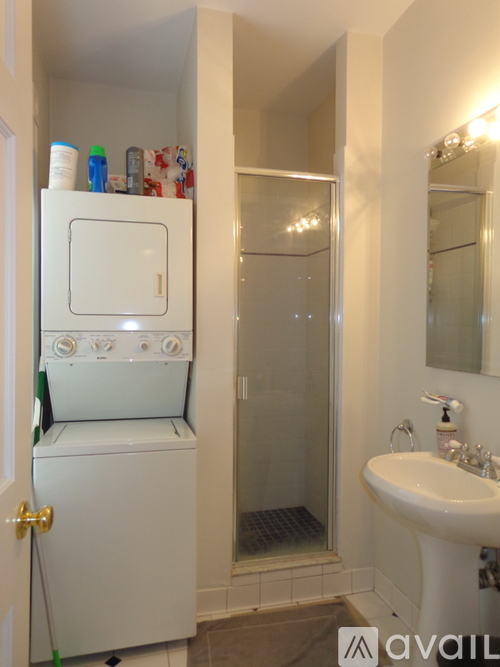 A small white refrigerator sits in a narrow bathroom next to a sink.