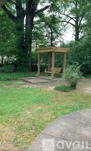 A wooden gazebo sits in a grassy area with trees in the background.