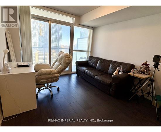 A living room with a brown couch and a chair with a computer desk in front of a window.