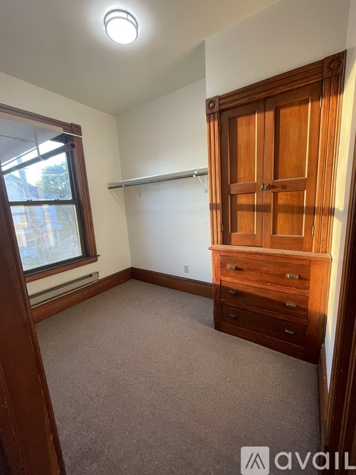 A bedroom with a window, a wooden wardrobe, and a carpeted floor.