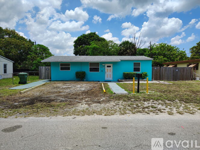 A blue house with a white door and windows is surrounded by a grassy area and a wooden fence.