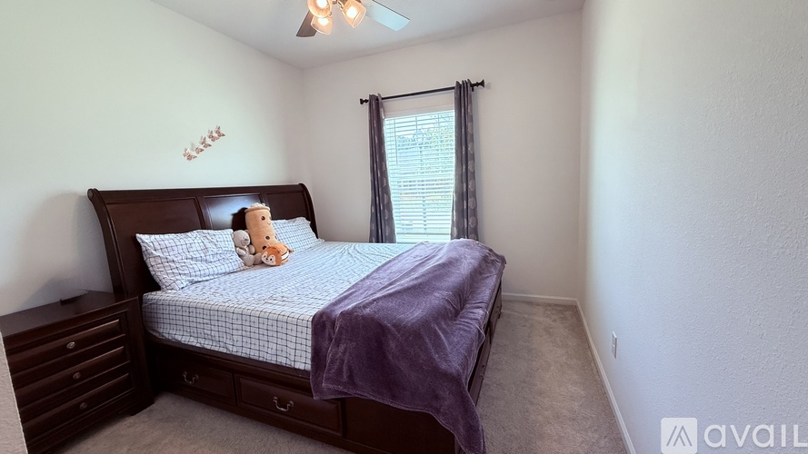 A bedroom with a bed, nightstand, and window with a teddy bear on the bed.