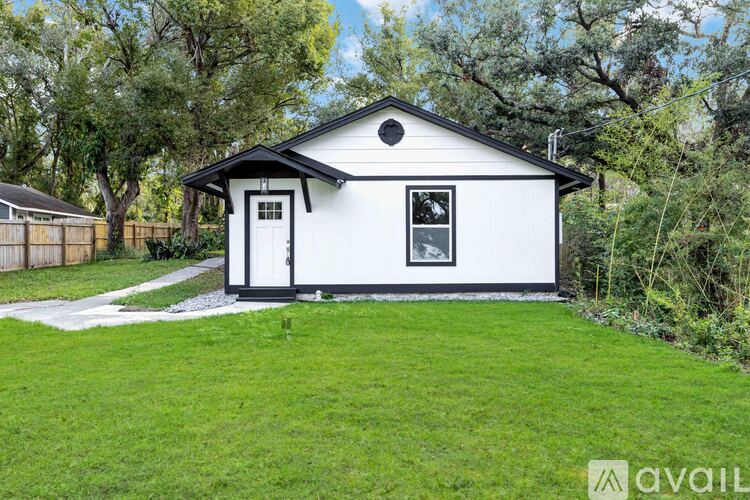 A small white house with a black roof and a black door is surrounded by green grass and trees.