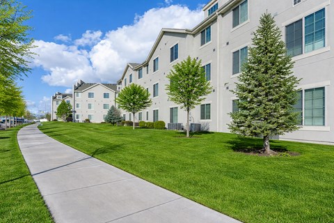 A long concrete walkway leads to a white building with green trees on the grassy lawn.