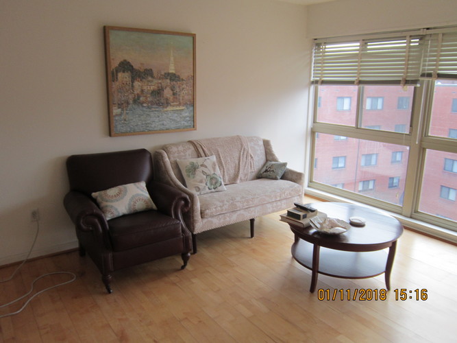 A brown couch sits in a living room with a painting on the wall and a window with blinds.