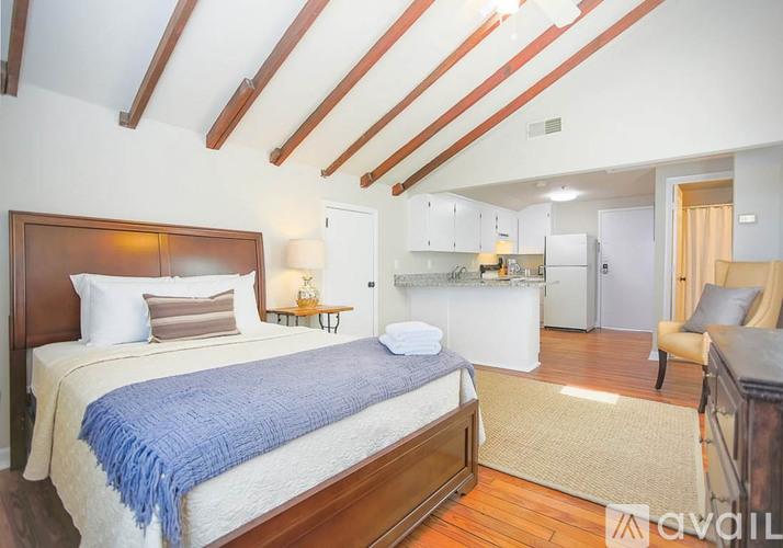 A bedroom with a bed, a chair, and a kitchen area in the background.