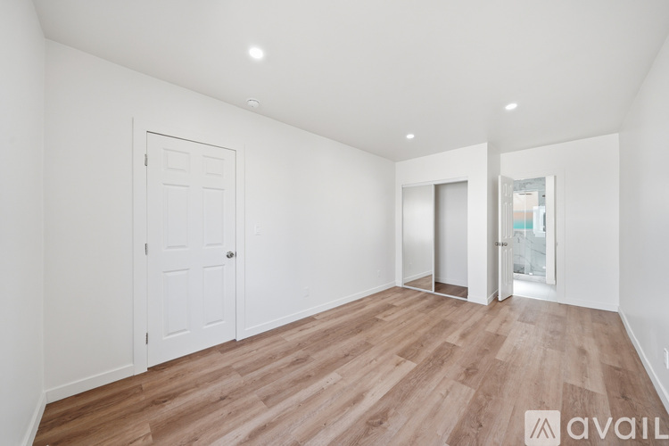 A room with wooden flooring and white walls, with a door on the left and a window on the right.