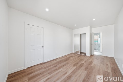 A room with wooden flooring and white walls, with a door on the left and a window on the right.