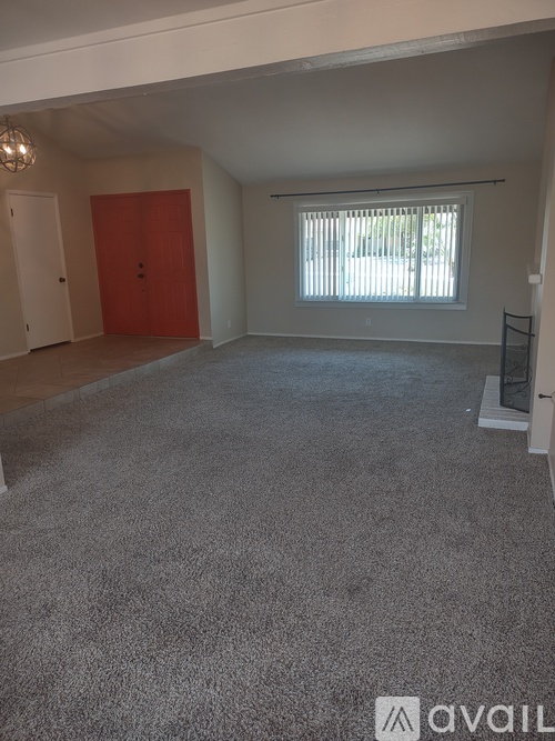 A room with a carpeted floor, a red door, and a window with blinds.