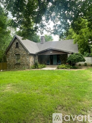 A house with a stone exterior and a grey roof is surrounded by a green lawn.