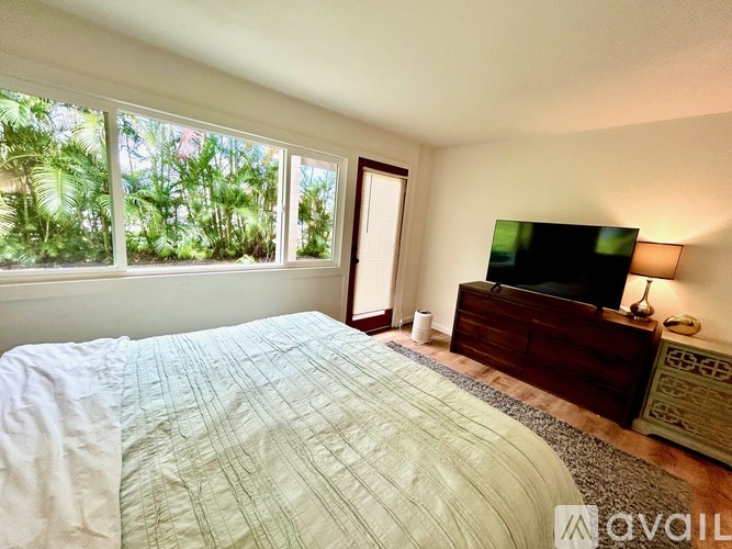 A bedroom with a large bed, a dresser with a flat screen TV on top, and a window with a view of greenery outside.