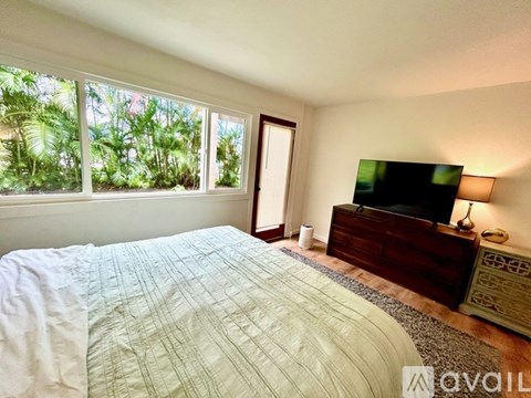 A bedroom with a large bed, a dresser with a flat screen TV on top, and a window with a view of greenery outside.