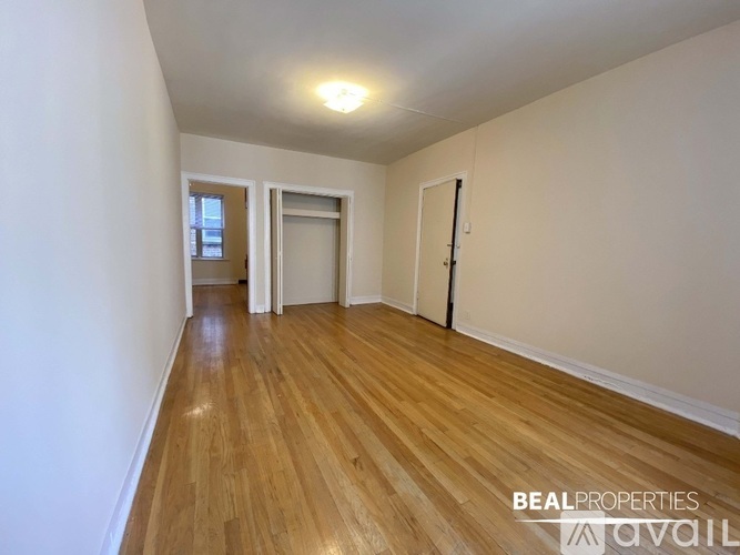 A room with wooden flooring and white walls, with a door on the right and a window on the left.
