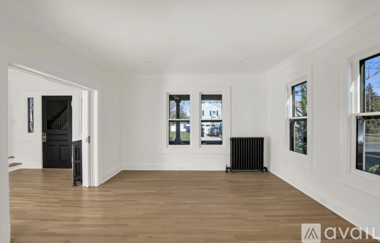 A spacious room with wooden floors and white walls, featuring a black door, a window with a view of trees, and a radiator on the floor.