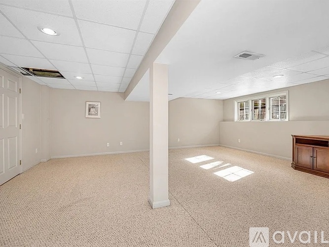 A spacious room with a carpeted floor, a door, a window with blinds, and a wooden cabinet.